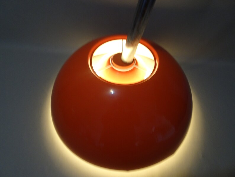 May include: A bright orange lampshade with a white interior and a silver metal stem. The lampshade is lit and casting a warm glow.