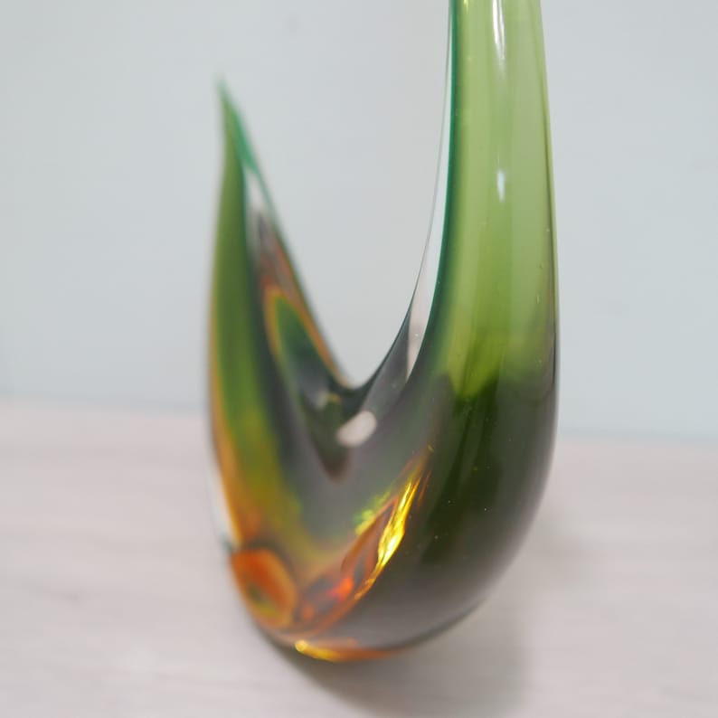 May include: A green and amber glass sculpture in the shape of a bird. The sculpture is abstract and has a smooth, glossy finish.