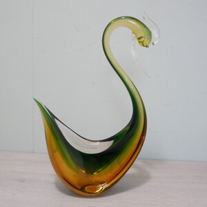 May include: A green and amber glass swan sculpture. The swan has a long neck and a curved body. The glass is clear and has a smooth finish.