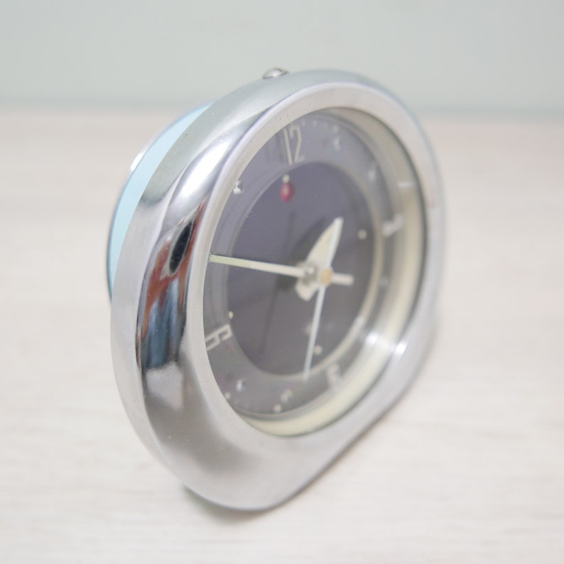 May include: A silver alarm clock with a black face and white hands. The clock has a blue and white band around the top.