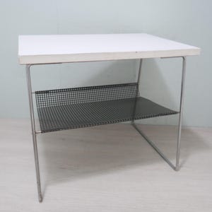 Può includere: Un tavolo in metallo bianco con un ripiano in filo metallico nero. Il tavolo ha un design semplice e moderno.