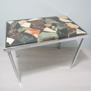 Peut inclure: Une table d'appoint avec un cadre en chrome et un plateau en verre noir orné d'une mosaïque de pierres colorées.