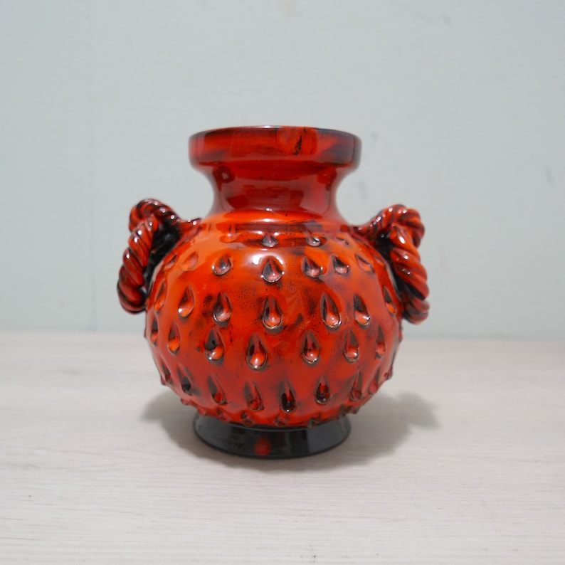 May include: A small, orange ceramic vase with two rope handles. The vase has a textured surface with small, raised dots.