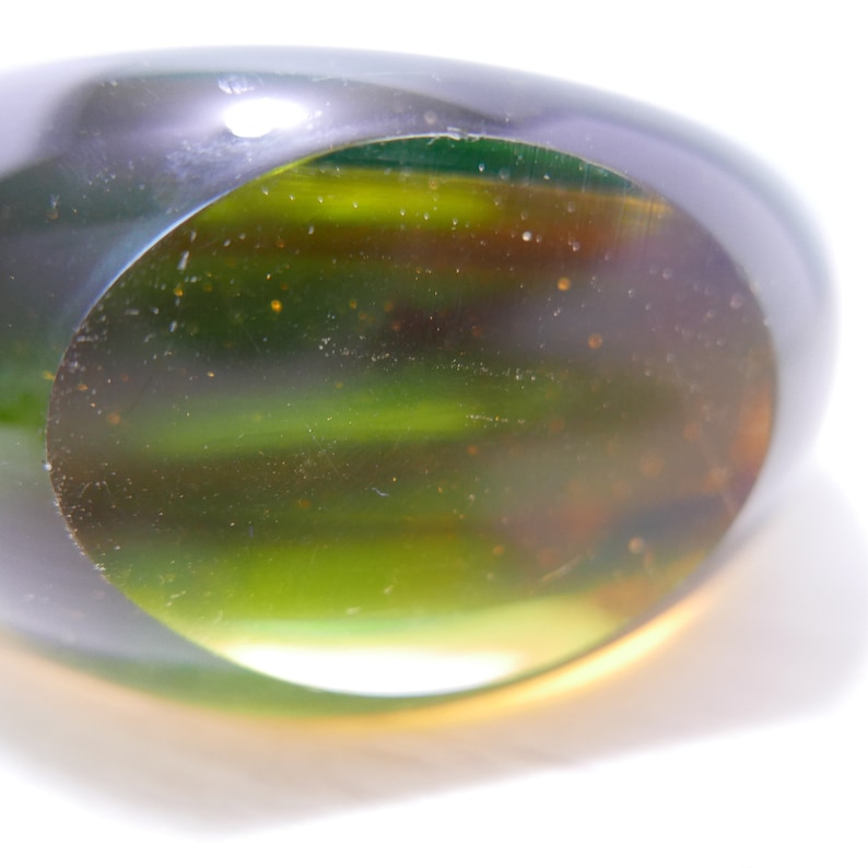 May include: A green glass paperweight with a round, clear center. The glass has a mottled, textured appearance.