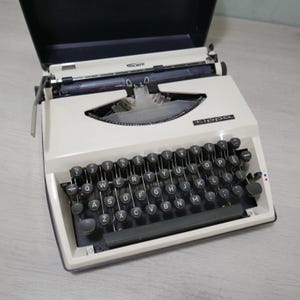 May include: Cream Triumph Tippa typewriter with black keys and a black case. The keyboard has a QWERTY layout. The typewriter has a metal carriage and a black top cover. The word "Triumph" is visible on the top.