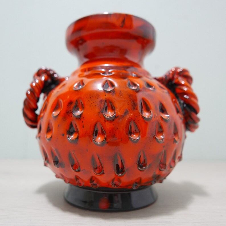 May include: Orange ceramic vase with a textured surface and two handles. The vase has a black base.