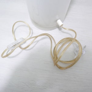 K&ouml;nnte beinhalten: Eine wei&szlig;e Lampe mit einem durchsichtigen Kabel und Stecker. Das Kabel ist aufgerollt und der Stecker ist sichtbar.