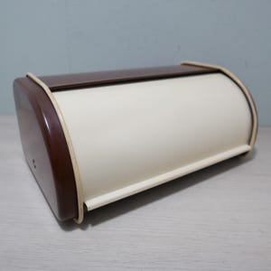 May include: A vintage bread box with a curved, roll-top design. The box features a cream-colored body with a dark brown lid and end panels. A thin, light-colored trim outlines the edges. The bread box is made of metal and is designed to store bread.
