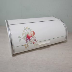 May include: White metal bread box with a roll-top lid and silver trim. The front features a colorful floral and fruit design, including grapes, apples, and berries. The bread box is designed to keep bread fresh and is a kitchen storage item.