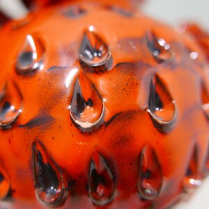 May include: Close-up of a bright orange ceramic object with a textured surface. The surface is covered in small, black, teardrop-shaped indentations.