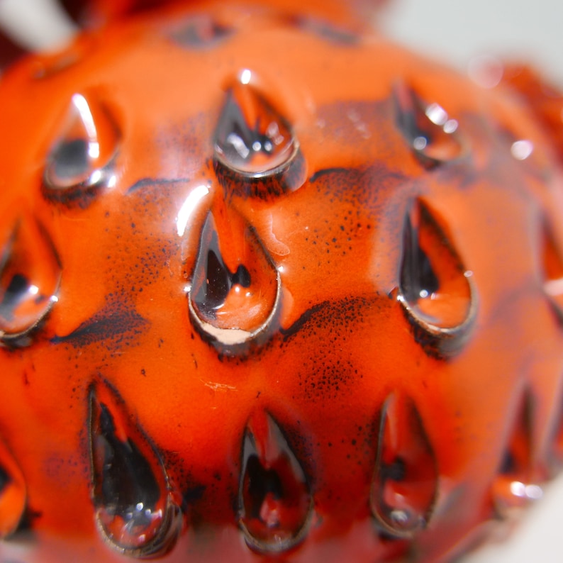 May include: Close-up of a bright orange ceramic object with a textured surface. The surface is covered in small, black, teardrop-shaped indentations.