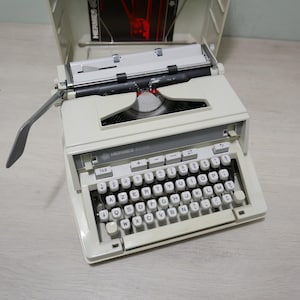 May include: An off-white Hermes 8008 typewriter with a full keyboard and a grey carriage return lever. The typewriter's case is open, revealing a red and black document. The keyboard has white keys with black lettering.