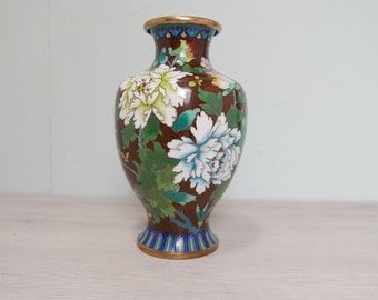May include: A brown cloisonné vase with a floral design. The vase is decorated with white, green, and blue flowers. The vase has a gold rim and base.