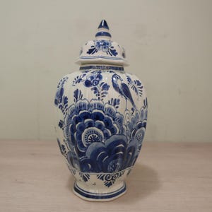 Op de afbeelding: Een wit en blauw keramieken pot met een deksel. De pot is versierd met blauwe bloemen- en vogelontwerpen. De pot heeft een bolvorm en een voetstuk. Het deksel heeft een kleine knop. De pot is waarschijnlijk een decoratief item.