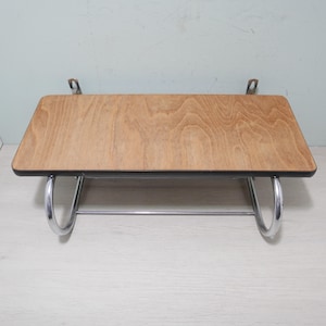 May include: A rectangular wooden shelf with a light brown finish and a black border. The shelf is supported by a chrome-coloured metal frame with curved legs and a horizontal bar. It is mounted on the wall with metal brackets.