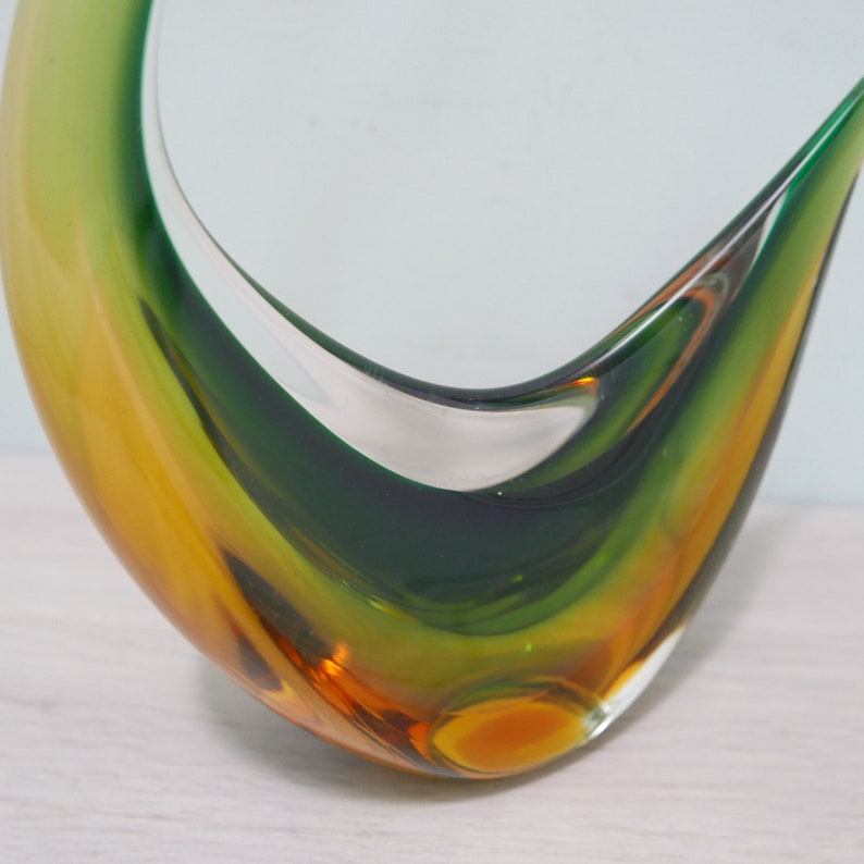 May include: A green and amber glass sculpture in the shape of a bird in flight. The sculpture is made of layered glass with a clear glass center.
