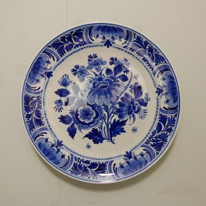 Könnte beinhalten: Ein runder, weißer Keramikteller mit blauem Blumendesign. Der Teller zeigt einen zentralen Blumenstrauß aus blauen Blumen und Blättern, umgeben von einem detaillierten blauen Rand. Das Design erinnert an Delfter Ware.