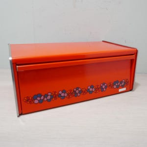 May include: A rectangular orange metal bread box with a hinged lid and silver trim. The front of the box features a decorative floral pattern in blue, pink, and orange. The bread box is a vintage kitchen storage item.