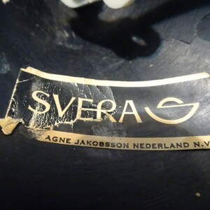 May include: A close-up of a black label with gold lettering that reads "SVERA AGNE JAKOBSSON NEDERLAND N.V."