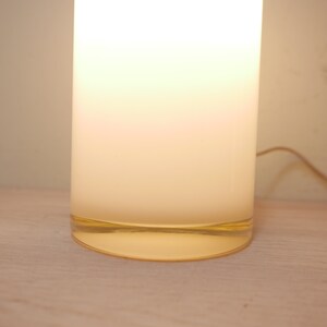 K&ouml;nnte beinhalten: Eine wei&szlig;e zylindrische Lampe mit einem klaren Glasfu&szlig;. Die Lampe ist eingeschaltet und strahlt ein warmes, gelbes Licht aus.