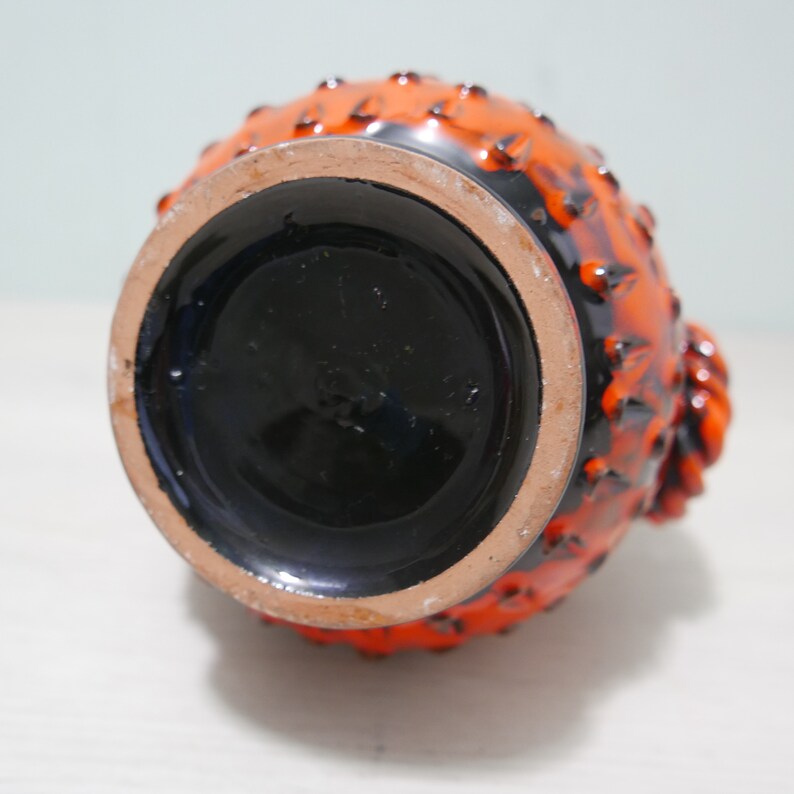 May include: Orange and black ceramic vase with a textured surface and a round base. The vase has a unique design with raised bumps and a textured pattern.