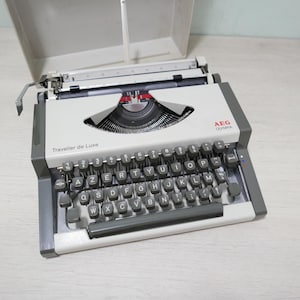 May include: A vintage AEG Olympia Traveller de Luxe typewriter in grey and white. The keyboard features black keys with white lettering. The typewriter's case is open, displaying the internal mechanisms. The text "Traveller de Luxe" is visible on the body.