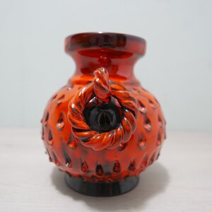 May include: Orange and black ceramic vase with a rope-like handle. The vase has a textured surface with small bumps.