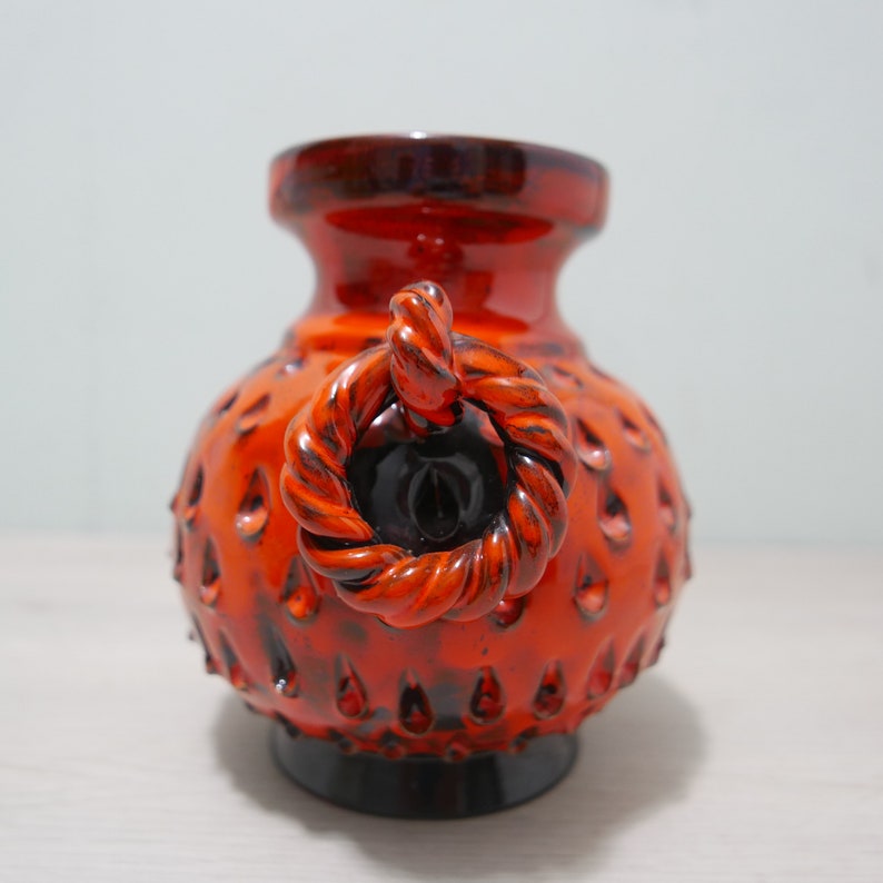 May include: Orange and black ceramic vase with a rope-like handle. The vase has a textured surface with small bumps.