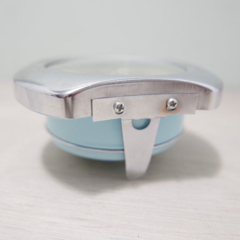 May include: A light blue metal alarm clock with a silver metal frame and two screws on the side.