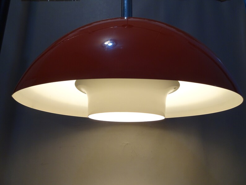 May include: A red and white ceiling lamp with a mushroom-shaped shade. The lamp is turned on and the light is shining through the white shade.