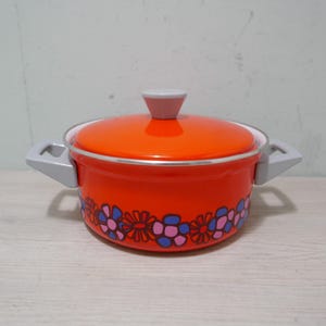 May include: An orange enamel cooking pot with a lid and two handles. The pot has a floral pattern around the base in blue, pink, and purple. The lid and handle are gray. The pot is approximately 8 inches in diameter.