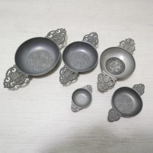 May include: A collection of pewter bowls with ornate handles. The bowls have a circular design in the center and decorative scrollwork on the handles. The bowls vary in size and are arranged on a white surface.