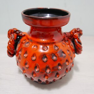May include: Orange ceramic vase with black accents and a textured surface. The vase has two rope-like handles on the sides.