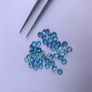 May include: A group of round, light blue gemstones, also known as topaz, scattered on a white surface. The gemstones are approximately 4mm in diameter.  AK Gems Beads is printed on the surface.