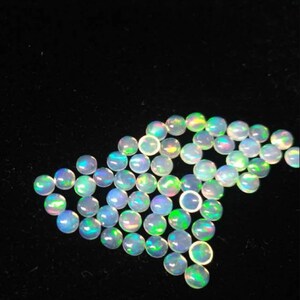 Può includere: Una collezione di cabochon di opale rotondi e iridescenti in varie tonalità di bianco, blu e verde. I cabochon sono disposti in un mucchio sciolto su uno sfondo nero.