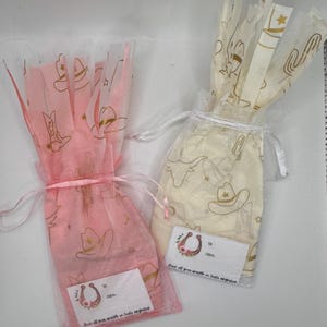 May include: Two small gift bags, one pink and one cream, are tied with ribbons. The bags are see-through and decorated with gold cowboy-themed illustrations. Each bag has a small white label with text and a horseshoe graphic.