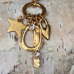 May include: A gold-toned charm necklace featuring a star, horseshoe, key, and heart pendant. The horseshoe and key charms are embellished with clear crystals. The necklace is displayed against a textured, light-colored background.