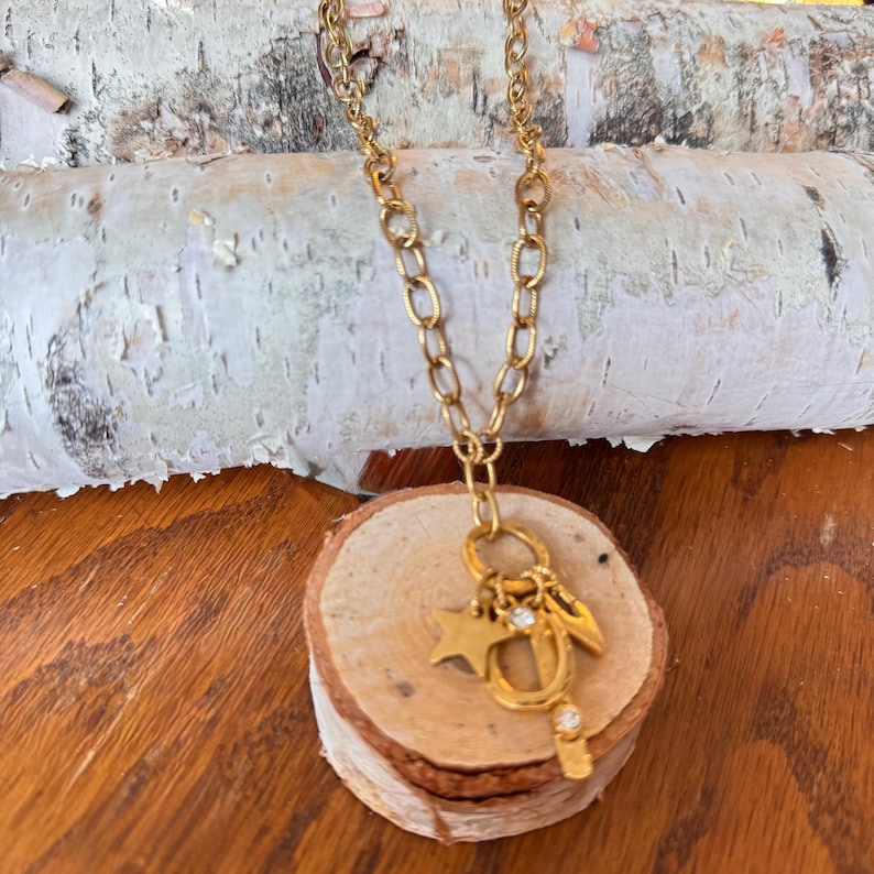 May include: A gold-colored necklace with a chain and multiple charms, including a star, a key, and a small crystal. The necklace is displayed on a small wooden slice, with a birch log in the background.