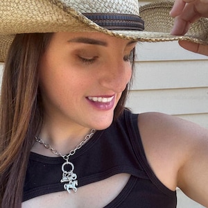 May include: Silver-toned necklace with a charm featuring a cowboy hat, horseshoe, and a horse. The necklace is worn with a black top and a straw cowboy hat with a brown band.