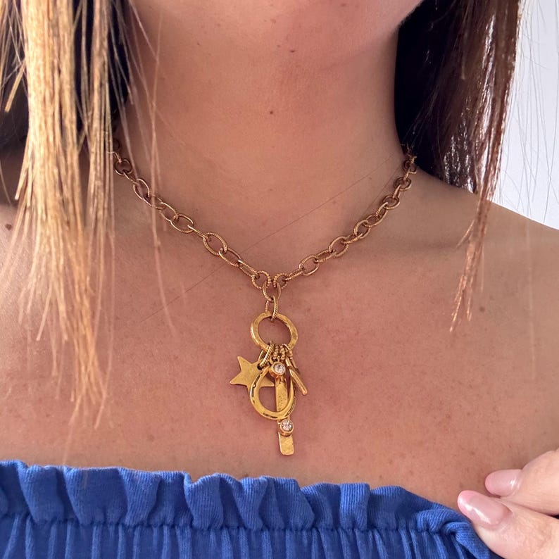 May include: Gold-toned necklace with a chunky chain and a pendant featuring a star, horseshoe, and other charms. The pendant also includes a small, clear gemstone. The necklace is worn against a skin tone and a blue top.