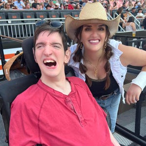 May include: Two people smiling at an outdoor event. One person is wearing a red Henley shirt and sunglasses on their head. The other person is wearing a straw cowboy hat, a white denim vest, and a black top. They are leaning against a metal railing.