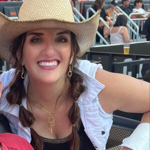 May include: A woman wearing a straw cowboy hat, gold earrings, and a gold necklace with a pendant. She is also wearing a white denim vest over a black top. The woman is smiling, and her hair is styled in braids.