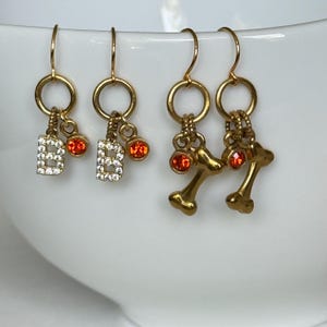 May include: Gold-tone earrings with a circular top and dangling charms. One pair features the letter "B" encrusted with clear crystals and a small orange gem. The other pair has bone-shaped charms and orange gems. The earrings are displayed against a white background.
