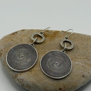 May include: A pair of silver earrings with a circular design. Each earring features a round, textured pendant with a spiral pattern, connected to a smaller, circular accent and a hook.