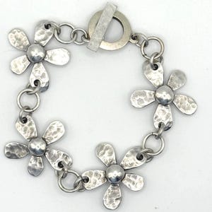 May include: A silver-toned metal bracelet featuring four flower-shaped charms with a hammered, textured finish. Each flower is connected by circular jump rings, and the piece is secured with a toggle clasp consisting of a ring and a bar.