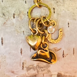 May include: A gold-toned charm cluster featuring a heart, cowboy hat, horseshoe, and a sparkling crystal. The charms are suspended from a circular ring, creating a rustic and charming aesthetic. The background is a light-colored, textured surface.