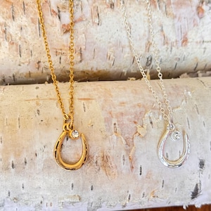 May include: Two delicate necklaces with horseshoe pendants and a single clear rhinestone accent. The necklaces are on a birch bark background. One necklace is gold and the other is silver.