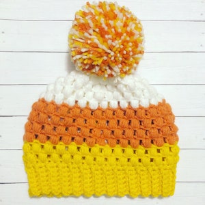 Può includere: Un cappello all'uncinetto a forma di mais caramellato. Il cappello è bianco, arancione e giallo. Ha un pon pon bianco in cima.