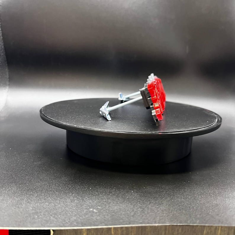 May include: A small, red and silver object with a distinctive design, possibly a decorative element or a component of a larger item. It is positioned on a black, circular platform, set against a dark background. The object's intricate details suggest a focus on aesthetics.