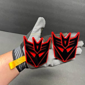 May include: Two black and red emblem badges with a geometric design. The badges are held in a gray work glove. The badges have a red outline and red accents within the black design.